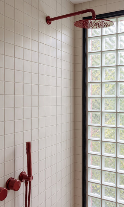 A modern bathroom corner with white square wall tiles, a dark red wall-mounted shower head and handheld shower attachment, and a glass block window with a black frame.