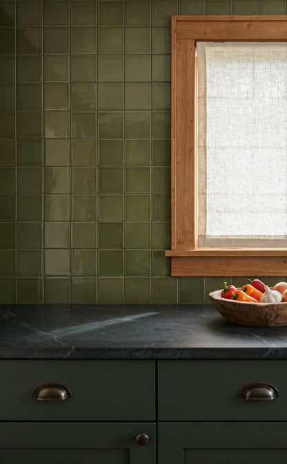 A kitchen scene with olive green subway tile backsplash, dark stone countertop, matching green cabinets, and a wood-framed window with a light curtain. A wooden bowl of fruit sits on the counter.