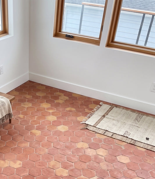 Hexagonal terracotta clay floor tiles in a room with white walls, wood-framed windows, and a woven rug on the floor.
