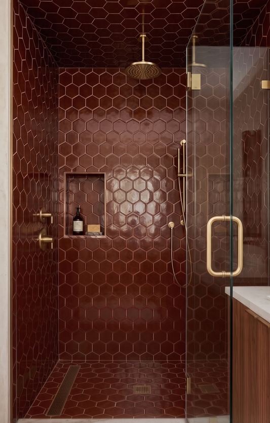 Deep reddish-brown hexagonal solid tile by Clay Imports covering a modern shower interior with gold fixtures and a glass door.