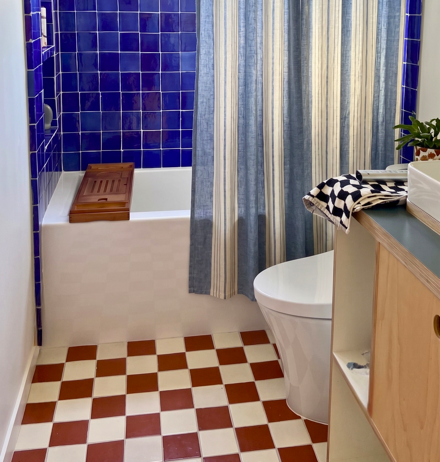 A retro-style bathroom featuring a white bathtub with a blue wooden bath tray, deep cobalt blue tiled walls, a blue and cream striped shower curtain, a white toilet, and a checkered floor in terracotta and cream.