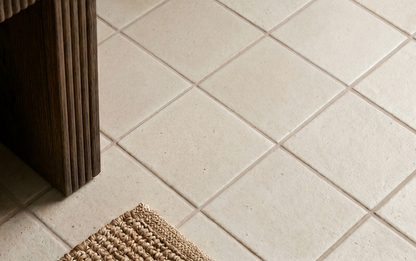 Cream-colored square solid tile with subtle speckled texture, laid in grid pattern, shown in lifestyle floor view with wooden furniture leg and woven mat corner.