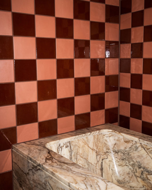 A close-up of a checkered tile wall in warm terracotta pink and dark brown, with a marble-look bathtub or sink edge in the foreground.