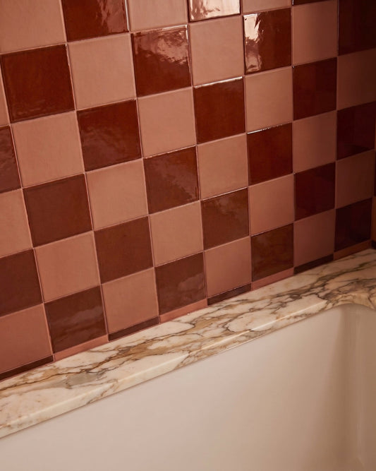 Glossy checkered wall tiles in alternating warm copper-rose and deep dark brown shades, installed above a white bathtub with a marble-look ledge.