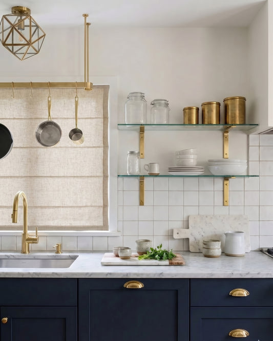 A styled kitchen interior featuring navy blue cabinets with brass handles, a white marble countertop, a gold faucet, glass shelves with canisters, and a beige window blind.