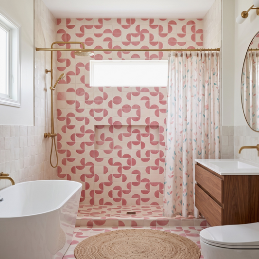 A modern bathroom interior featuring a bold pink and beige geometric patterned tile wall, a matching shower curtain, a freestanding white bathtub, a wooden vanity, and a round woven rug.