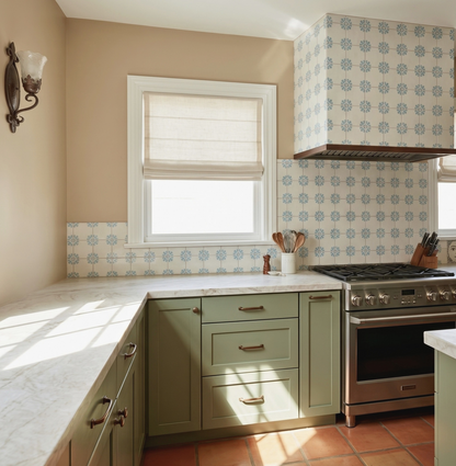 A kitchen interior with sage green cabinets, white marble countertops, a blue and white patterned tile backsplash, terracotta tile flooring, a stainless steel gas range, and a window with a white blind.