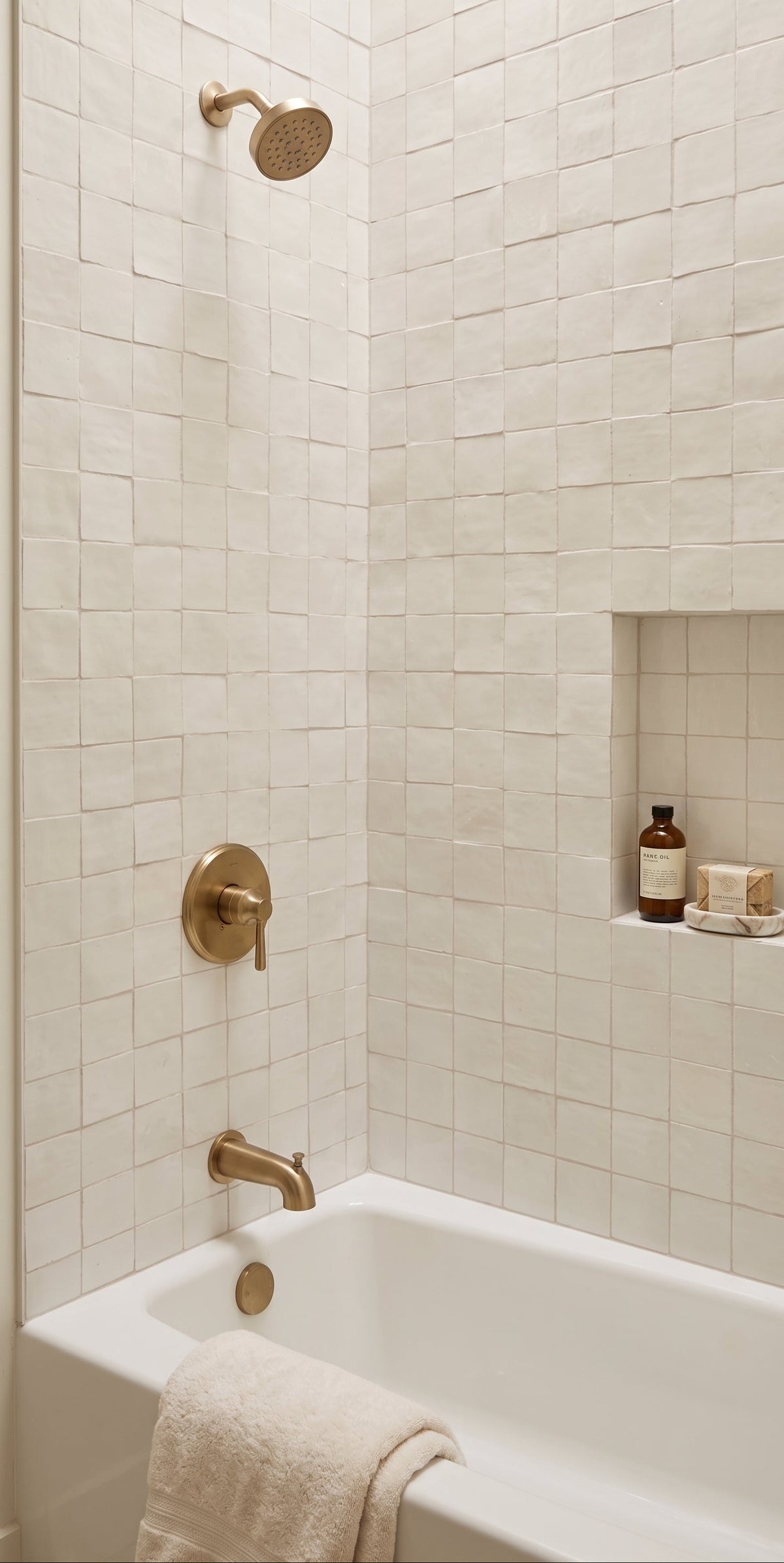 A white bathtub installed in a tiled bathroom corner with a recessed shelf holding small bottles and a folded towel draped over the tub edge. The wall and niche are covered in small off-white rectangular tiles, and the fixtures are a warm brass/gold tone.