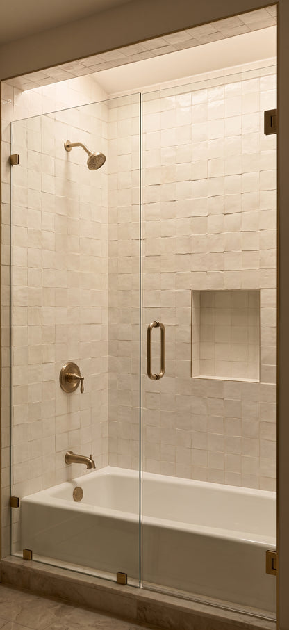 A modern frameless glass shower door installed in a walk-in shower enclosure with a white tiled wall, built-in niche, and white bathtub. The fixtures have a brushed brass/gold finish.