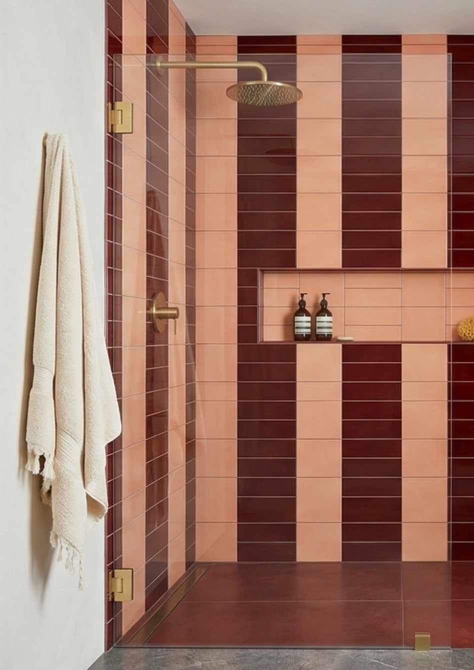 Vertical striped shower tiles by Clay Imports in burgundy and wildflower, featuring brass fixtures and a recessed niche.