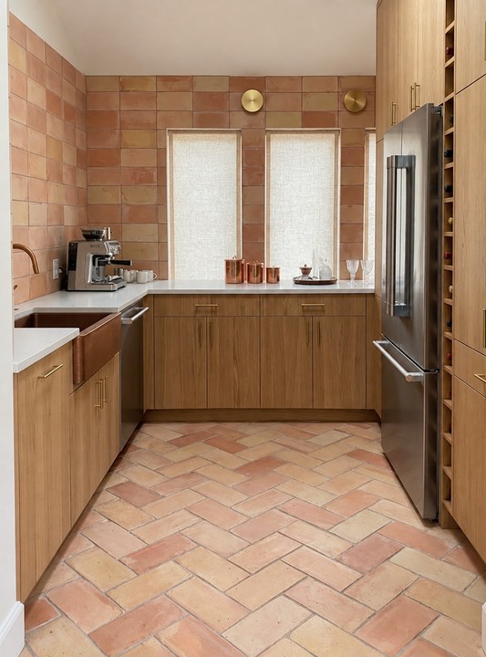 A modern kitchen interior with light wood cabinets, terracotta tile backsplash and floor, white countertops, two windows with sheer curtains, and a stainless steel refrigerator.