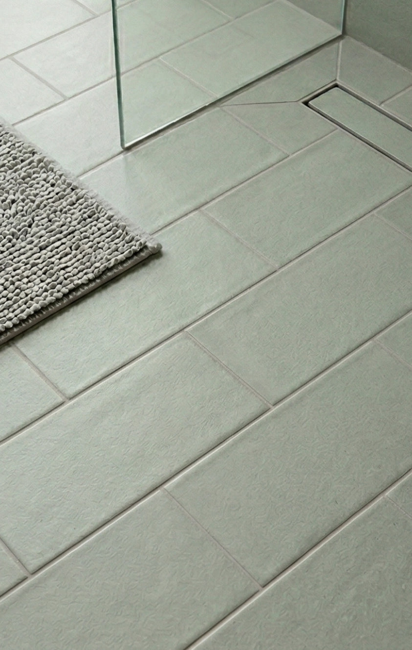 Light green rectangular textured clay tiles by Clay Imports laid diagonally on a bathroom floor with a glass shower screen, linear drain, and grey bath mat.