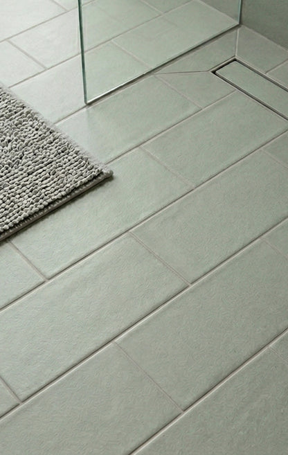 Light green rectangular textured clay tiles by Clay Imports laid diagonally on a bathroom floor with a glass shower screen, linear drain, and grey bath mat.