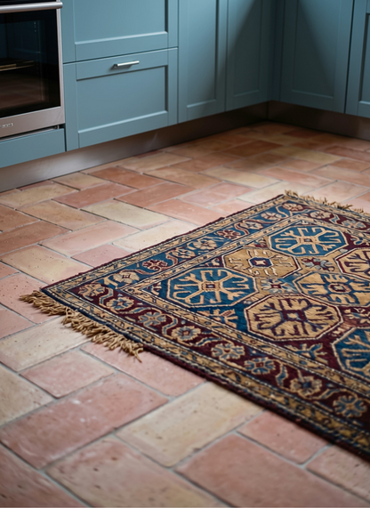 A traditional patterned area rug with blue, red, and beige geometric motifs lies on a terracotta tile kitchen floor in front of light blue cabinets.