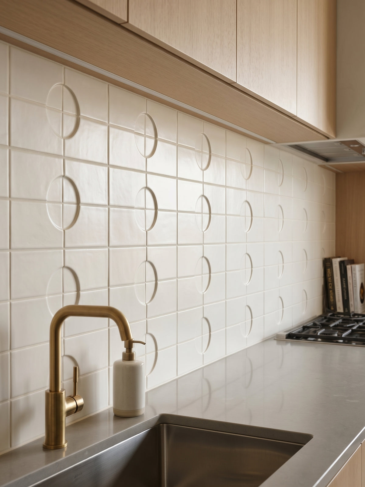 A sleek modern kitchen features a brass faucet and soap dispenser against an ivory tiled backsplash with debossed geometric circles.