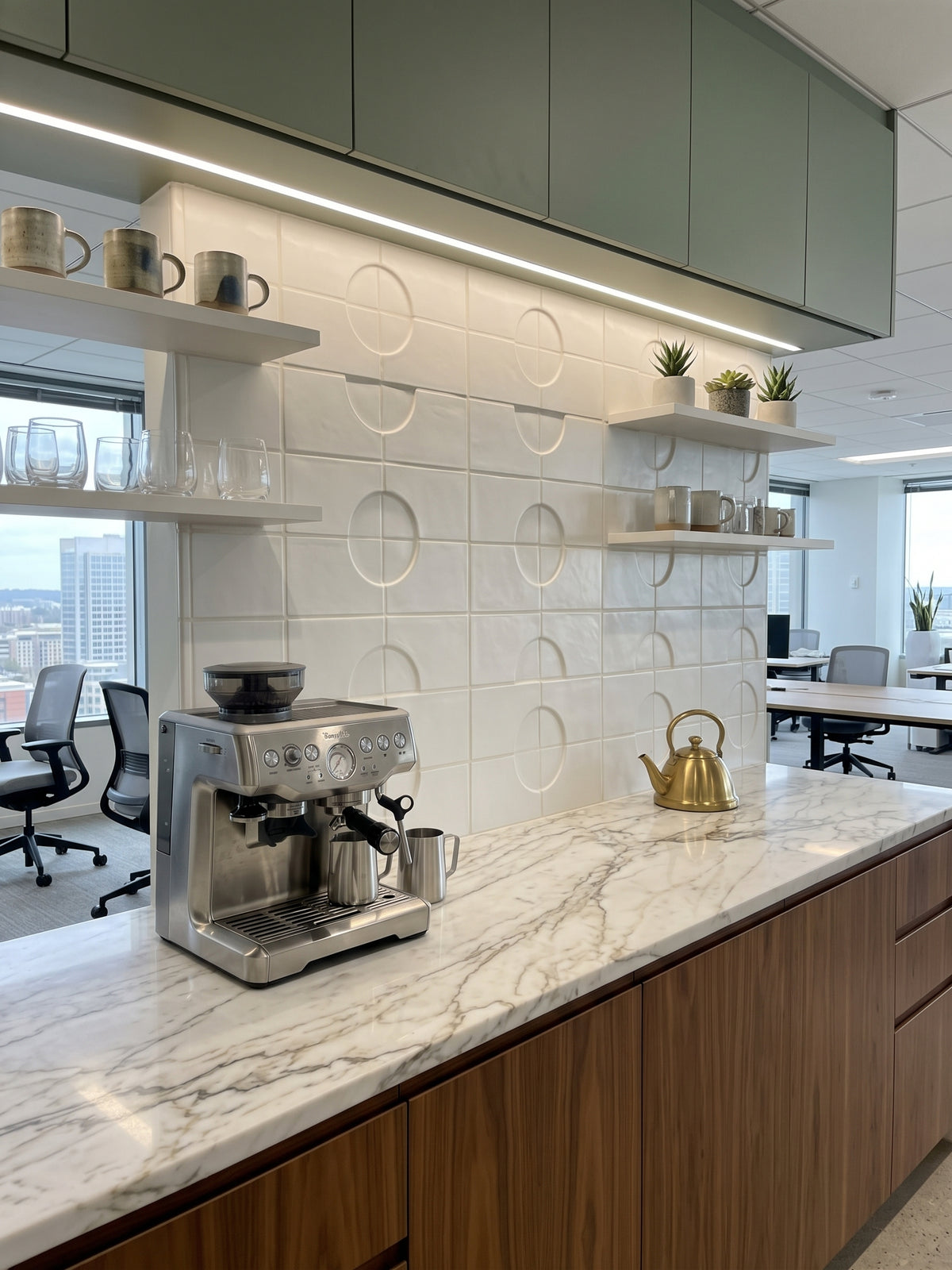 A stainless steel espresso machine and a gold kettle sit on a white marble countertop in a modern office breakroom featuring textured white tile walls and wooden cabinets.