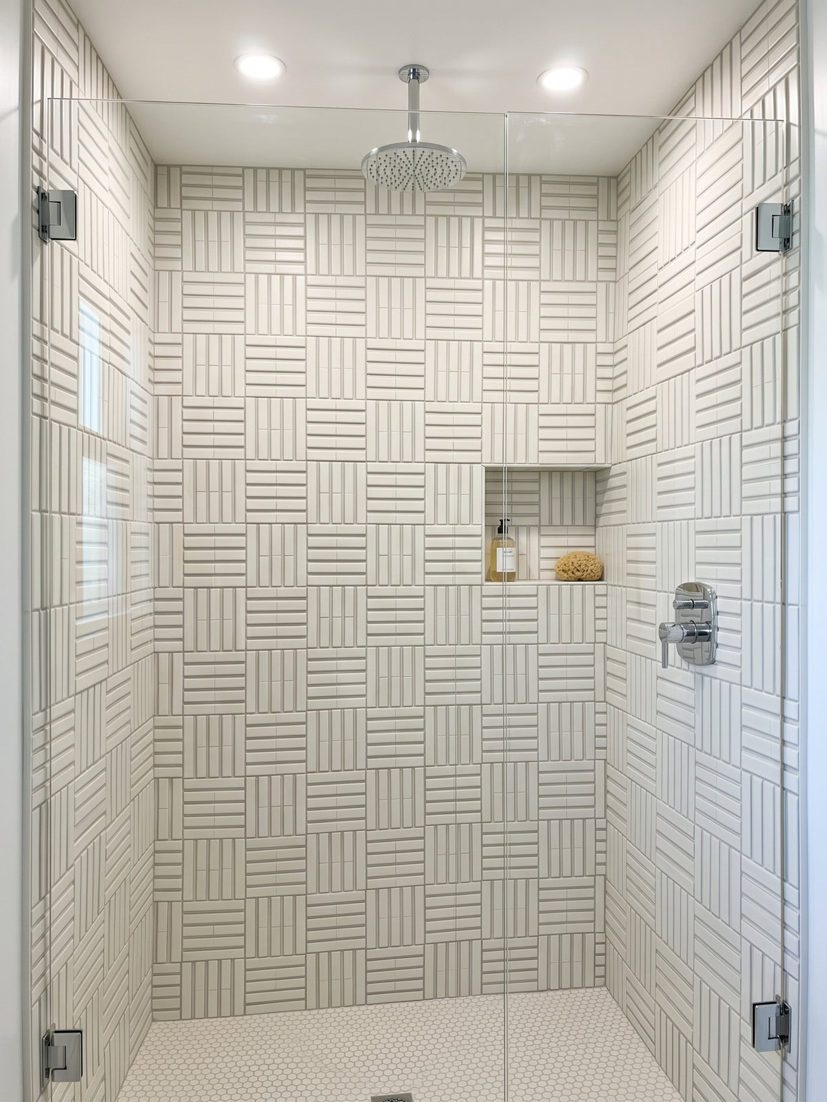 A modern walk-in shower features textured white basketweave tile walls, a rain showerhead, and a built-in shower niche with soap and a loofah.