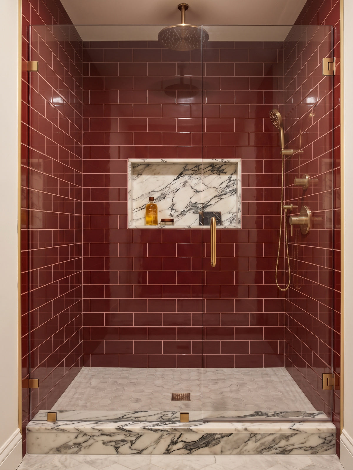 A modern walk-in shower enclosure with deep glossy red subway tile walls, a recessed marble niche, and a marble-look floor. The shower features matte brass fixtures, including a rain showerhead, handheld shower, and controls, along with a clear glass door.