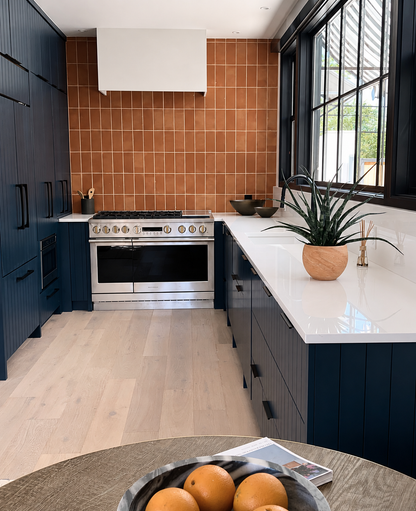 A modern kitchen interior with dark navy blue cabinets, a white countertop, a terracotta tiled backsplash, and a stainless steel gas range. A potted plant sits on the counter, and a bowl of oranges is visible in the foreground on a dining table.