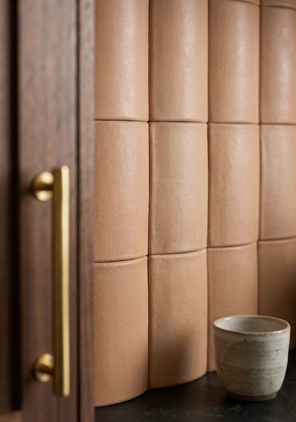 Warm beige curved clay tiles by Clay Imports forming a textured, dimensional wall, beside a wooden cabinet and ceramic cup.