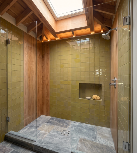 A modern skylight shower with olive green wall tiles, a gray stone floor, and warm wood paneling and ceiling beams.