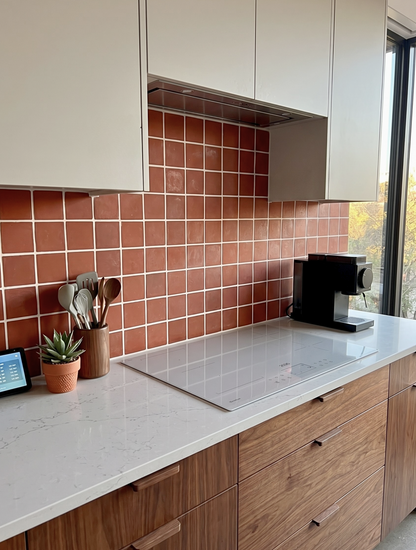 A modern kitchen corner with terracotta square wall tiles, a white countertop, wood cabinetry, a built-in cooktop, and a black coffee machine near a window.