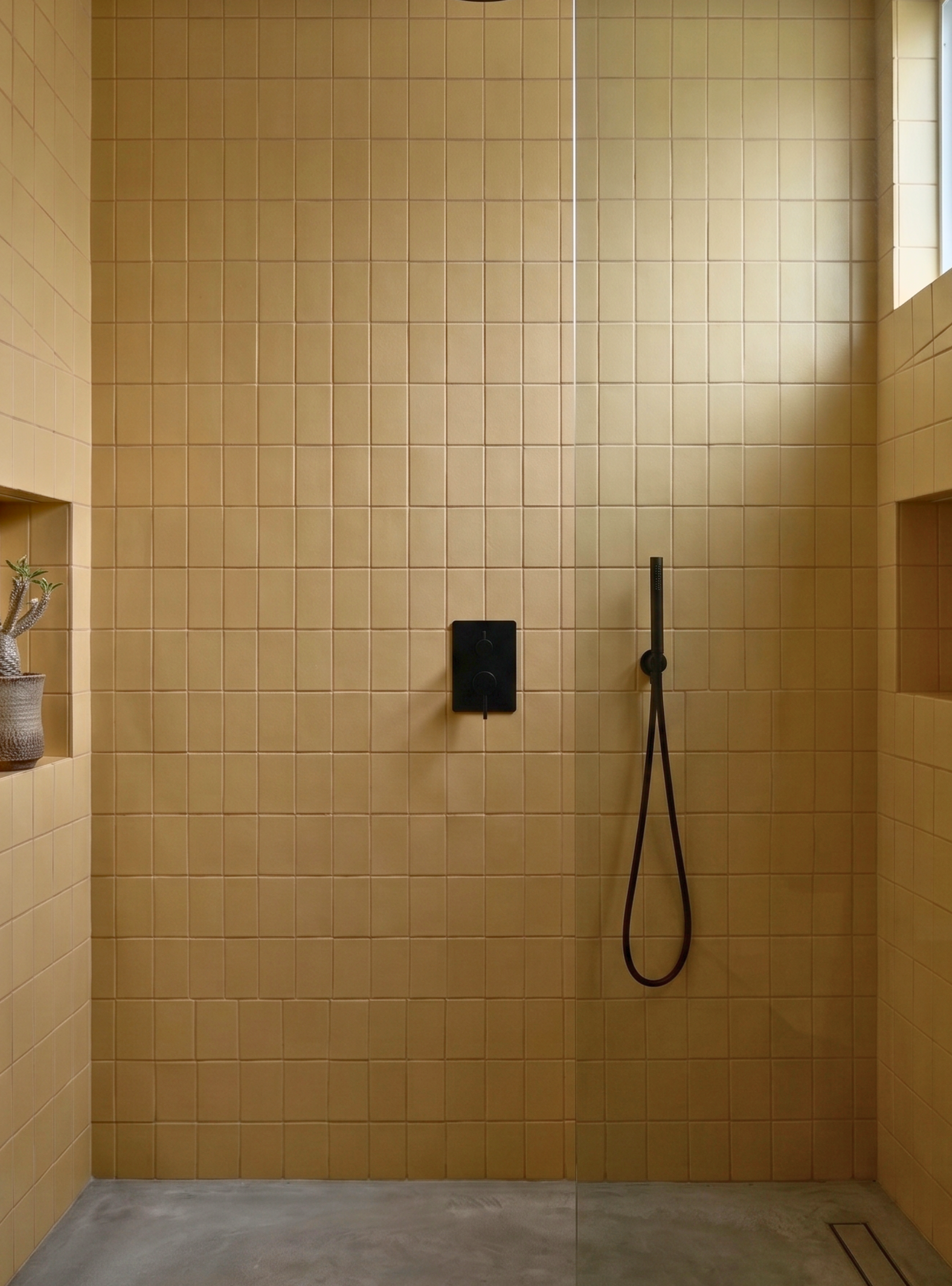 A modern shower area with warm beige square wall tiles, a black wall-mounted control panel, and a black handheld shower hose. A clear glass door is visible on the right, and a small recessed shelf with a plant appears on the left.