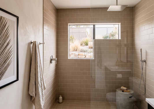 A modern bathroom shower area with beige subway tile walls and floor, a clear glass shower screen, a window looking out to a garden, a wall-mounted shower head and handheld shower, and a towel hanging on a wall rack.
