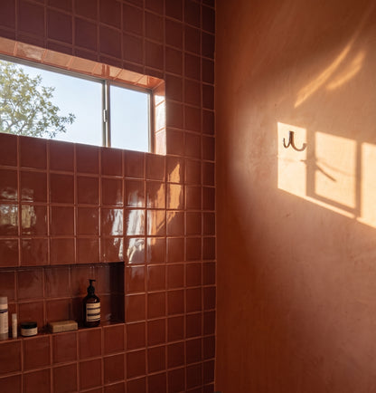 Sunlight streams through a bathroom window, casting shadows across glossy terracotta square tiles and a matching peach-toned wall with a metal robe hook.
