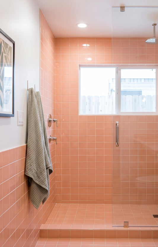 A modern bathroom features a walk-in shower with glossy peach square tiles, a glass door, and nickel fixtures, complemented by sage green waffle-weave towels hanging on the adjacent wall.