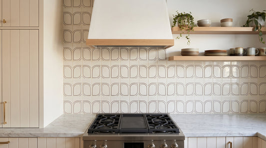 A kitchen backsplash featuring square tiles with a repeating arched geometric pattern in warm beige and cream tones, paired with a white range hood and a stainless steel gas cooktop.