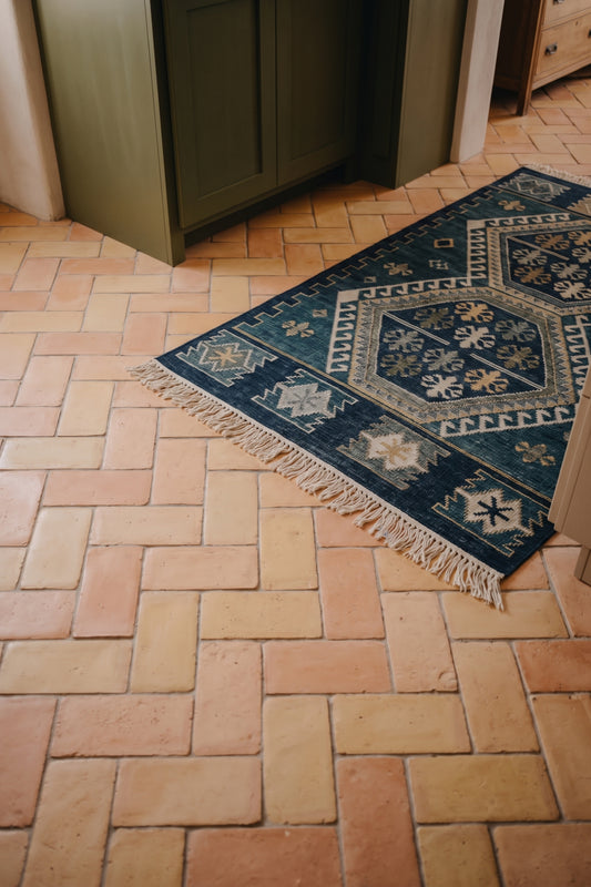 A dark navy blue area rug with cream and beige geometric patterns and fringe, styled on a terracotta tile floor in a home interior.