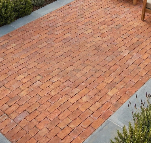 Reddish-orange terracotta Baked Paver 5x9.5 by Clay Imports Pavers, laid in a herringbone pattern patio bordered by grey stone and plants.