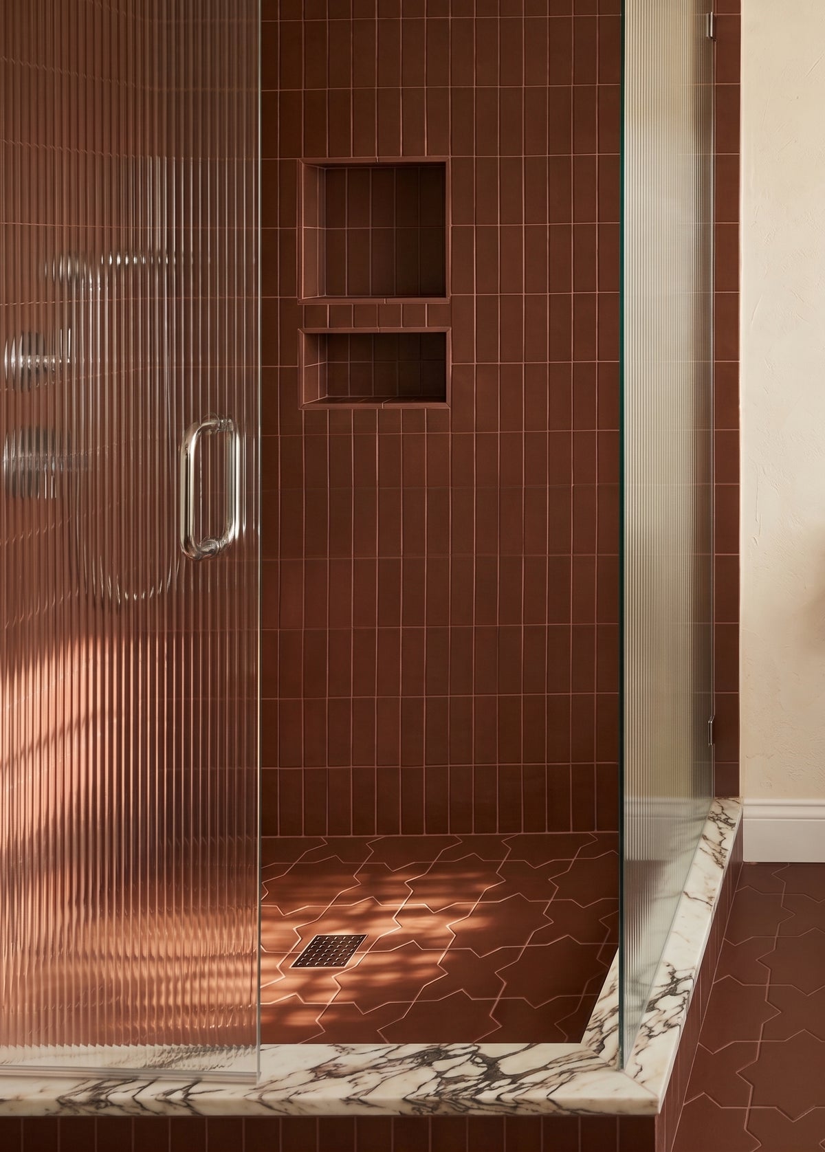A modern walk-in shower with a glass door featuring vertical ribbed texture, set against a wall of glossy brown rectangular tiles with built-in niche shelves and a matching brown tiled floor. The shower base has a white and gray veined marble pattern.