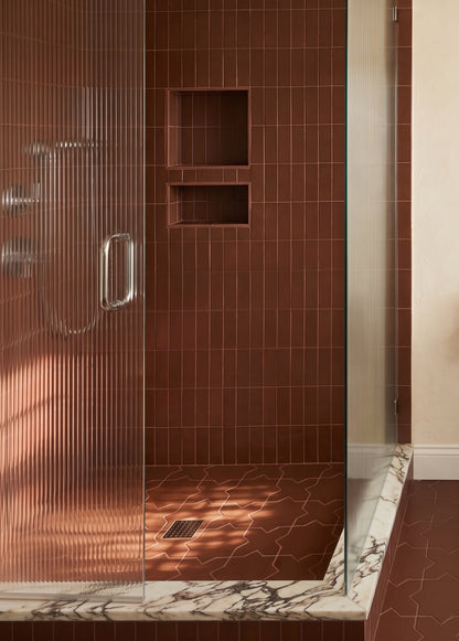 A modern walk-in shower with a glass door featuring vertical ribbed texture, set against a wall of glossy brown rectangular tiles with built-in niche shelves and a matching brown tiled floor. The shower base has a white and gray veined marble pattern.