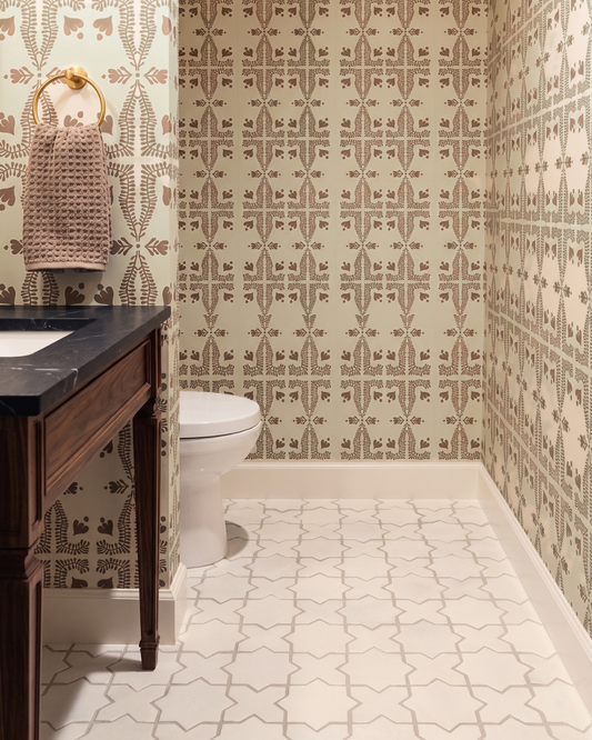 A bathroom interior featuring patterned wallpaper in beige and brown, a white toilet, a dark wood vanity with a black countertop, and a patterned white tile floor.