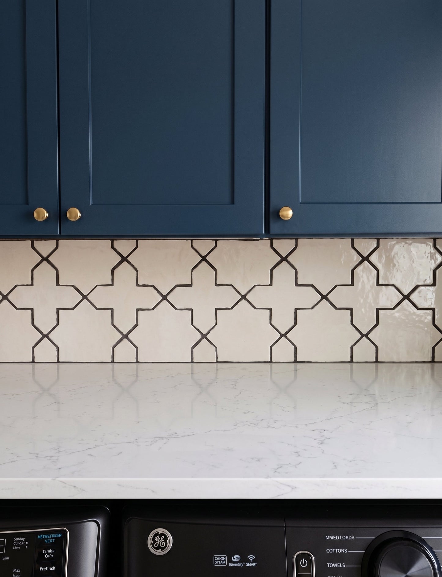 A close-up of a kitchen backsplash featuring cream-colored ceramic tiles with a black geometric trellis pattern, set beneath dark blue cabinets and a white marble countertop.