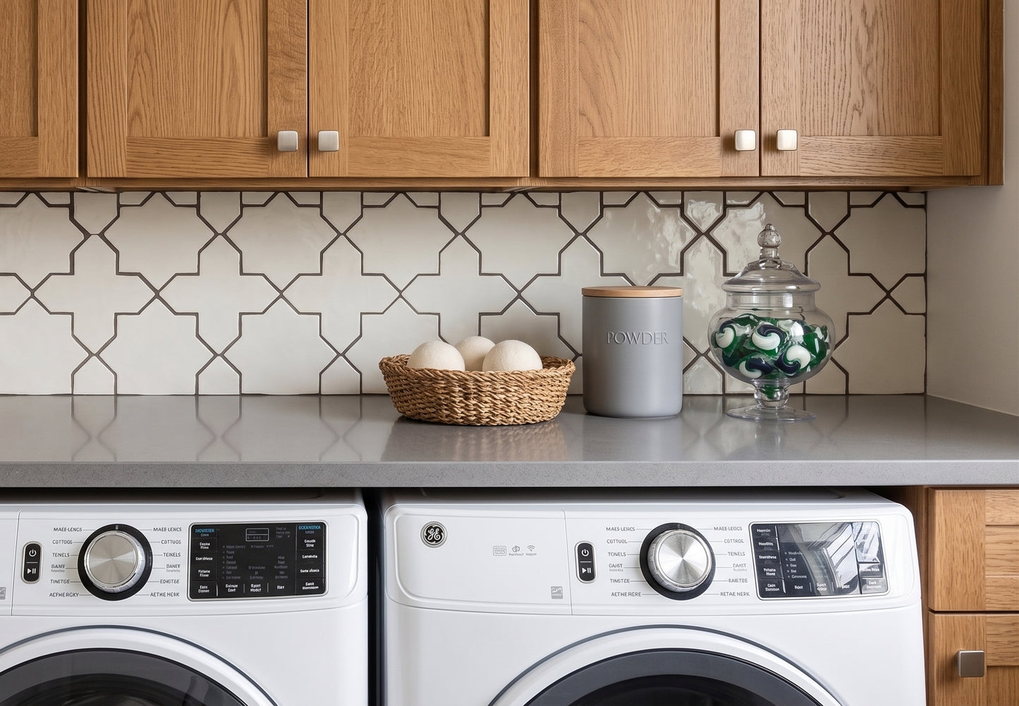 A modern laundry room corner featuring a gray countertop with a patterned tile backsplash, wooden cabinets, a basket of laundry eggs, a gray canister, and a glass jar of green-and-white laundry pods, with a white front-loading washing machine and dryer below.