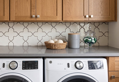 A modern laundry room corner featuring a gray countertop with a patterned tile backsplash, wooden cabinets, a basket of laundry eggs, a gray canister, and a glass jar of green-and-white laundry pods, with a white front-loading washing machine and dryer below.