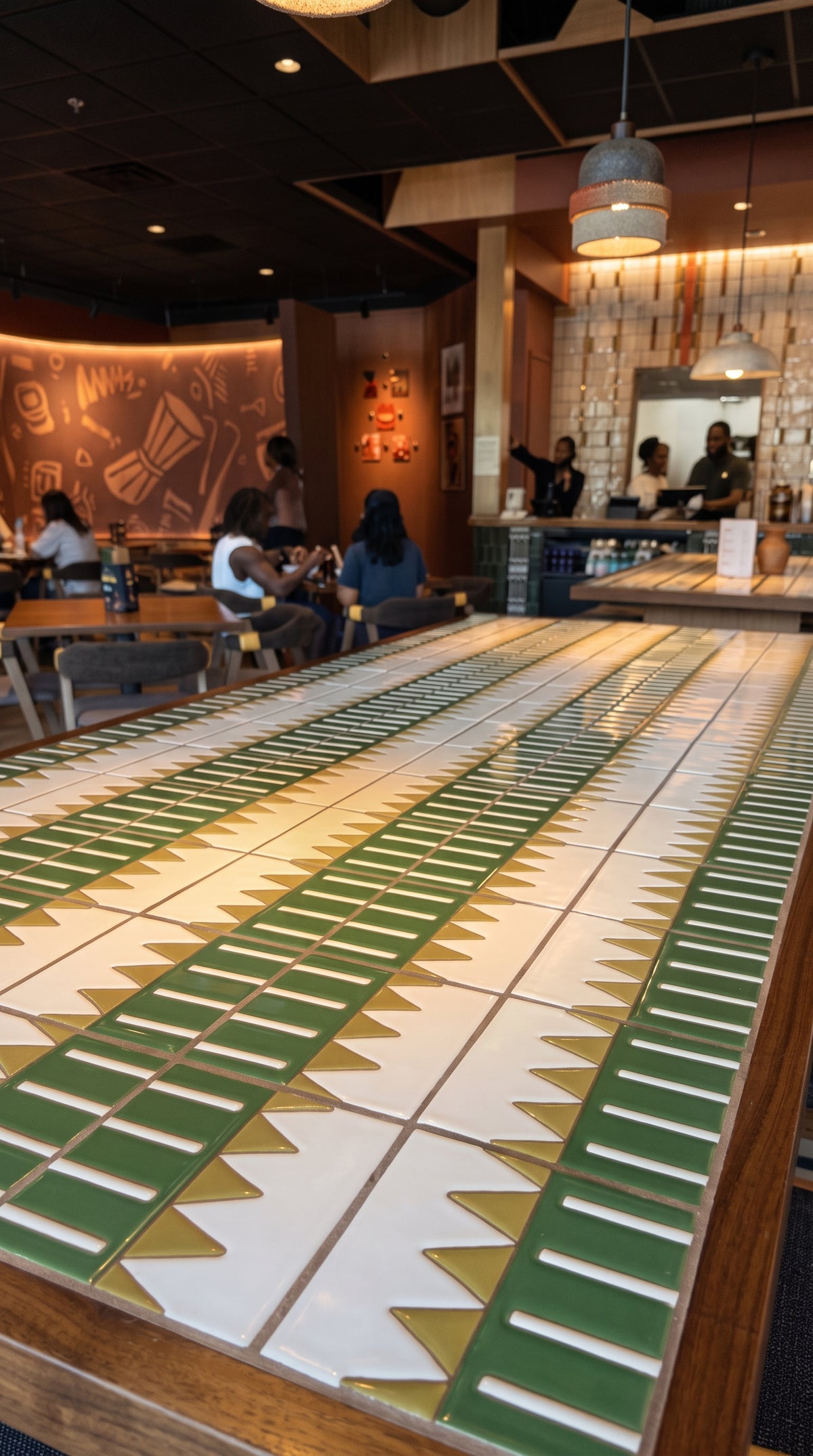A long dining table featuring green and white geometric tile patterns sits in the foreground of a modern, warmly lit restaurant interior.
