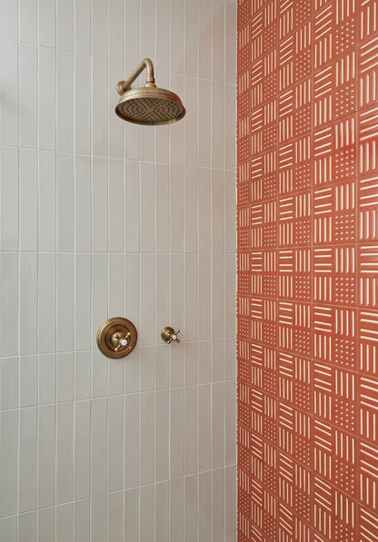 A close-up of a shower area with a round brass shower head and matching control valve on a white tiled wall, next to a terracotta-colored wall with a repeating geometric pattern.