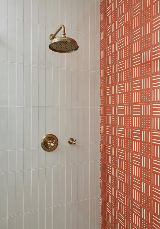 A close-up of a shower area with a round brass shower head and matching control valve on a white tiled wall, next to a terracotta-colored wall with a repeating geometric pattern.