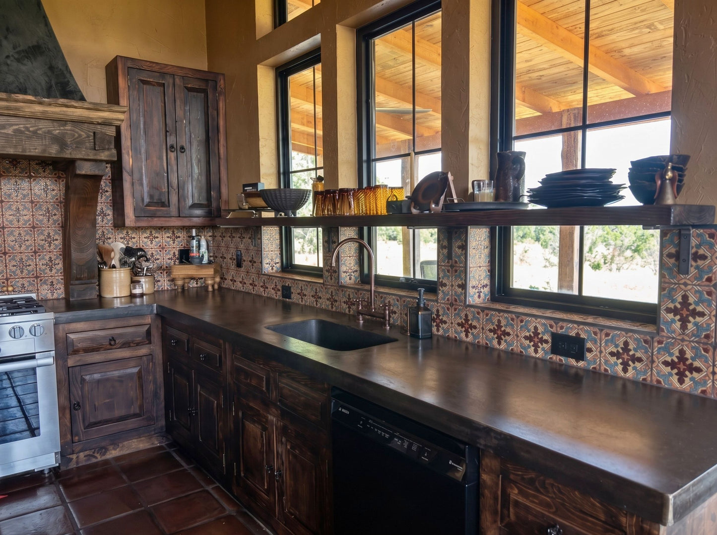 A rustic kitchen interior with dark wood cabinets, a black countertop sink, patterned tile backsplash, and large windows looking out onto a wooden deck.
