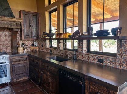 A rustic kitchen interior with dark wood cabinets, a black countertop sink, patterned tile backsplash, and large windows looking out onto a wooden deck.