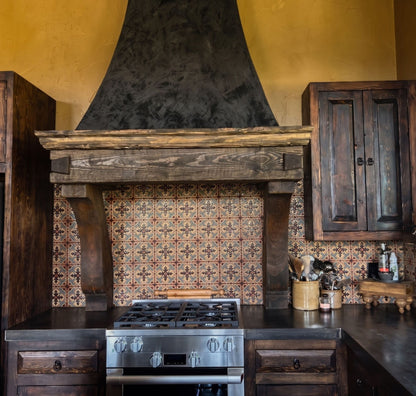 Rustic kitchen scene with a large dark metal range hood mounted above a wooden mantel and patterned tile backsplash, with dark wood cabinetry and a stainless steel gas range below. 