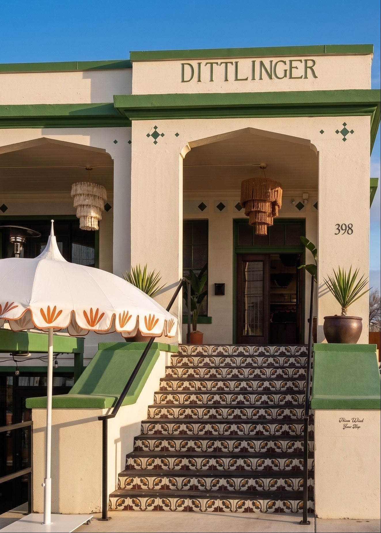 The exterior of the historic Dittlinger Hotel in Los Angeles, featuring a cream-colored stucco facade, green trim, patterned tile steps, a white patio umbrella with orange leaf motifs, and a clear blue sky.