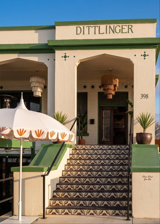 The exterior of the historic Dittlinger Hotel in Los Angeles, featuring a cream-colored stucco facade, green trim, patterned tile steps, a white patio umbrella with orange leaf motifs, and a clear blue sky.