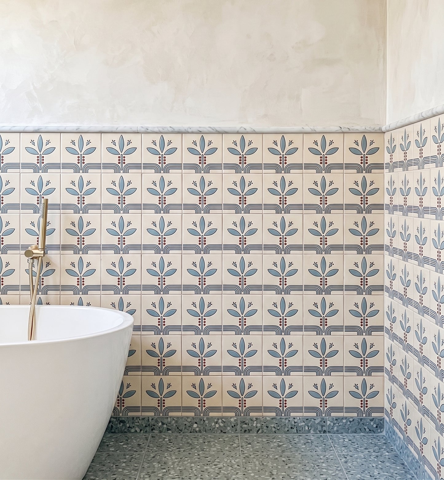 A bathroom corner showing patterned ceramic wall tiles with a repeating blue and gray floral motif on a warm cream background, a white freestanding tub edge, and a gray speckled floor.