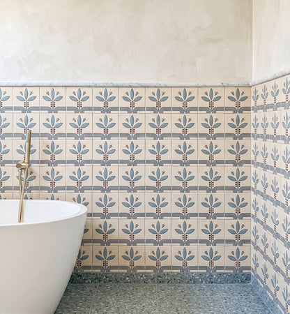 A bathroom corner showing patterned ceramic wall tiles with a repeating blue and gray floral motif on a warm cream background, a white freestanding tub edge, and a gray speckled floor.