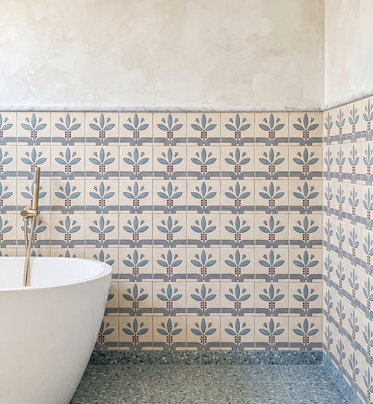 A bathroom corner showing patterned ceramic wall tiles with a repeating blue and gray floral motif on a warm cream background, a white freestanding tub edge, and a gray speckled floor.
